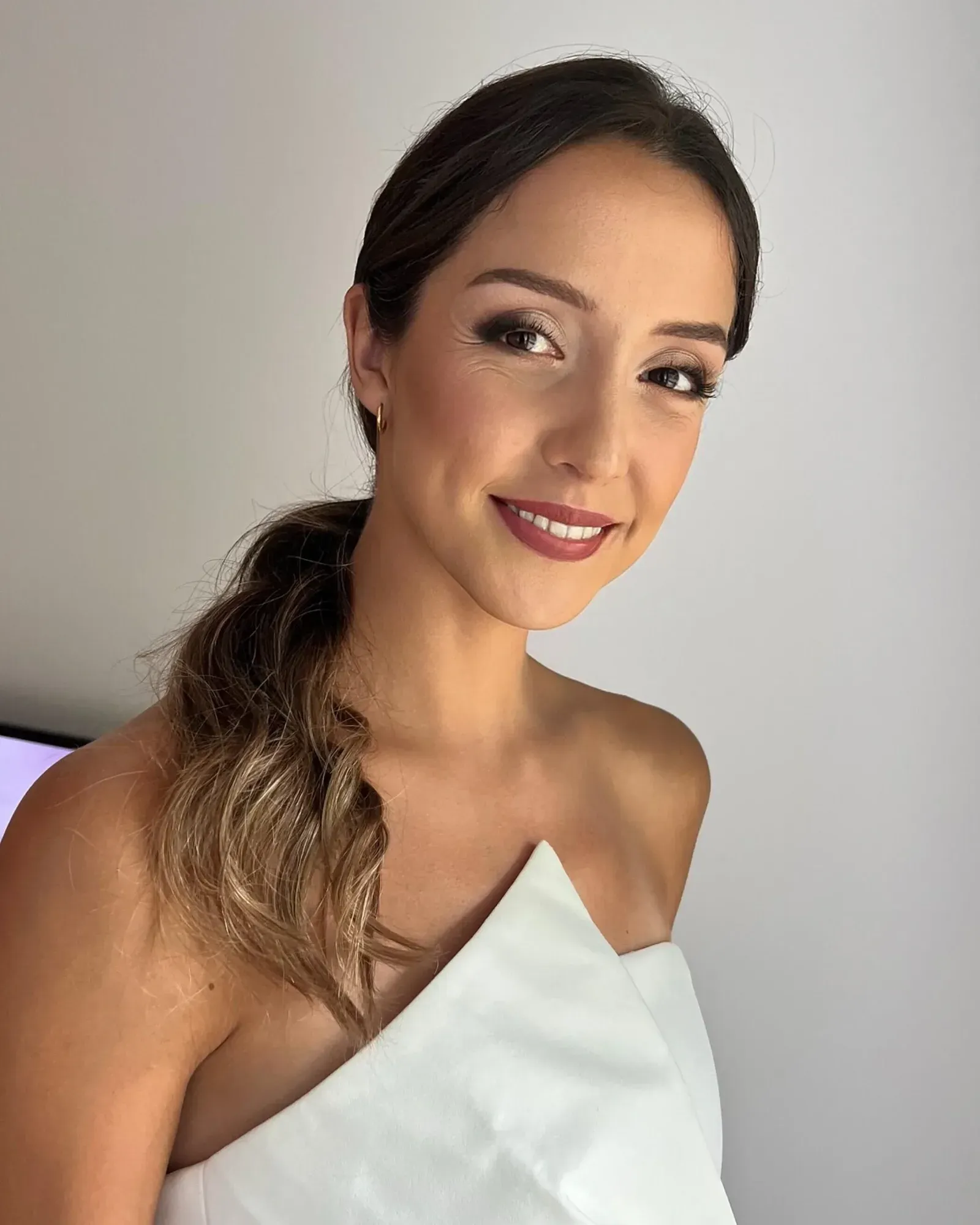 Une femme aux longs cheveux bruns ondulés et au teint clair, souriante et maquillée (rouge à lèvres, ombre à paupières et mascara), vêtue d'un haut blanc sans bretelles, sur un fond blanc uni.