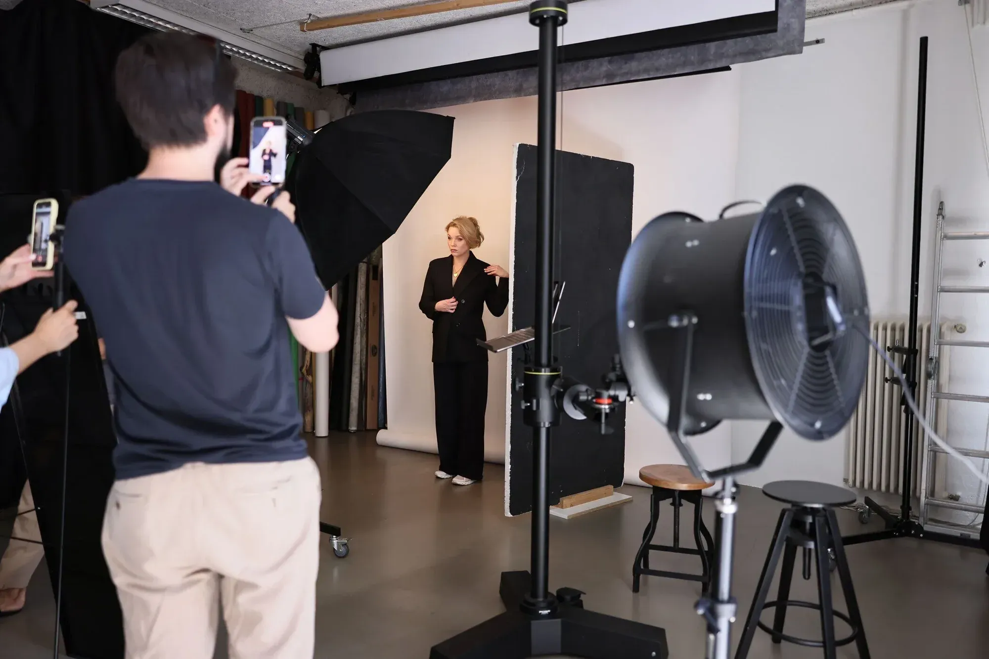 Une femme en tailleur noir pose devant une toile de fond photographique lors d'une séance de photos dans un studio avec du matériel photographique.
