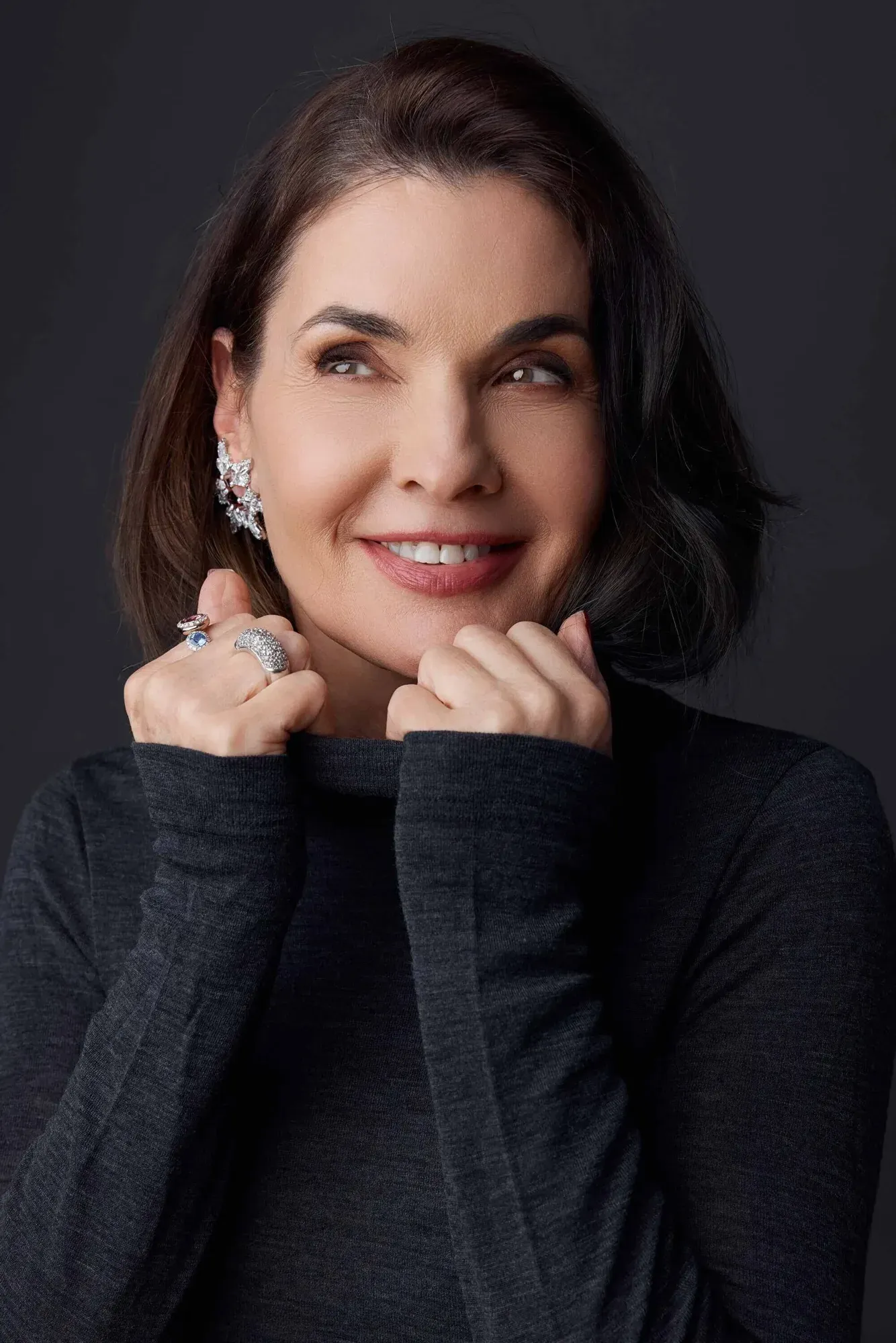 Une femme aux cheveux foncés coiffés en boucles souples, vêtue d'un haut noir à manches longues et portant de gros bijoux en argent, souriante et se touchant le menton, sur un fond sombre.