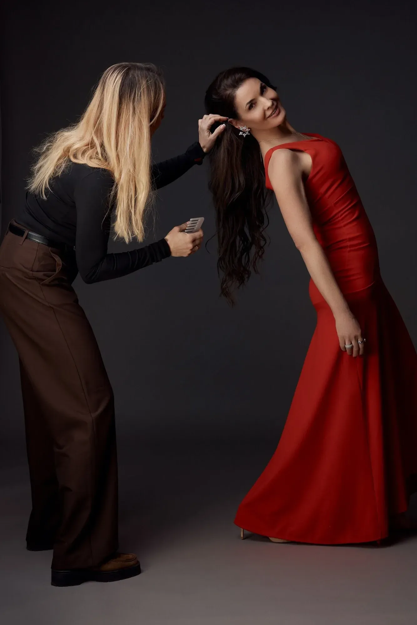 Une femme en robe rouge aux longs cheveux foncés sourit, tandis qu'une autre femme aux cheveux blonds, vêtue d'un haut noir et d'un pantalon marron, se touche les cheveux et tient un peigne.