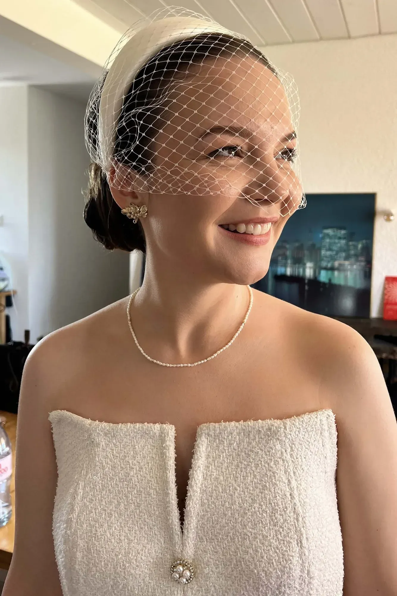 Une femme habillée pour un mariage, portant une robe blanche sans bretelles, un collier, des boucles d'oreilles assorties et un voile cage, avec un bouquet épinglé sur le devant de sa robe. Elle a les cheveux bruns courts et sourit en regardant vers sa droite, avec un arrière-plan