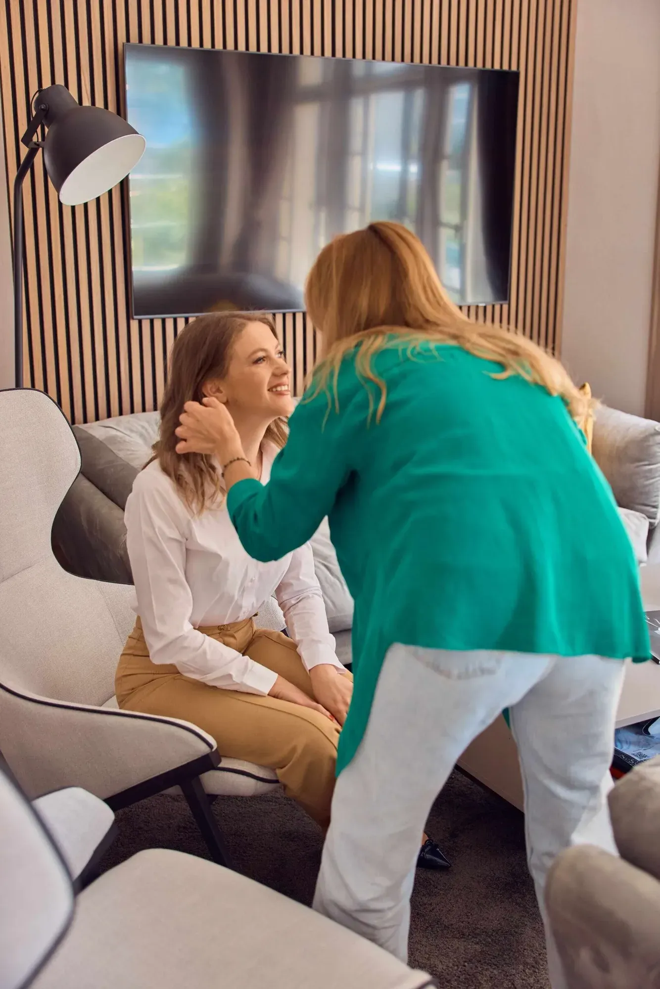 Une femme en chemise turquoise touche délicatement le visage d'une autre femme assise sur une chaise et souriante dans un salon avec une grande télévision sur le mur derrière elles.