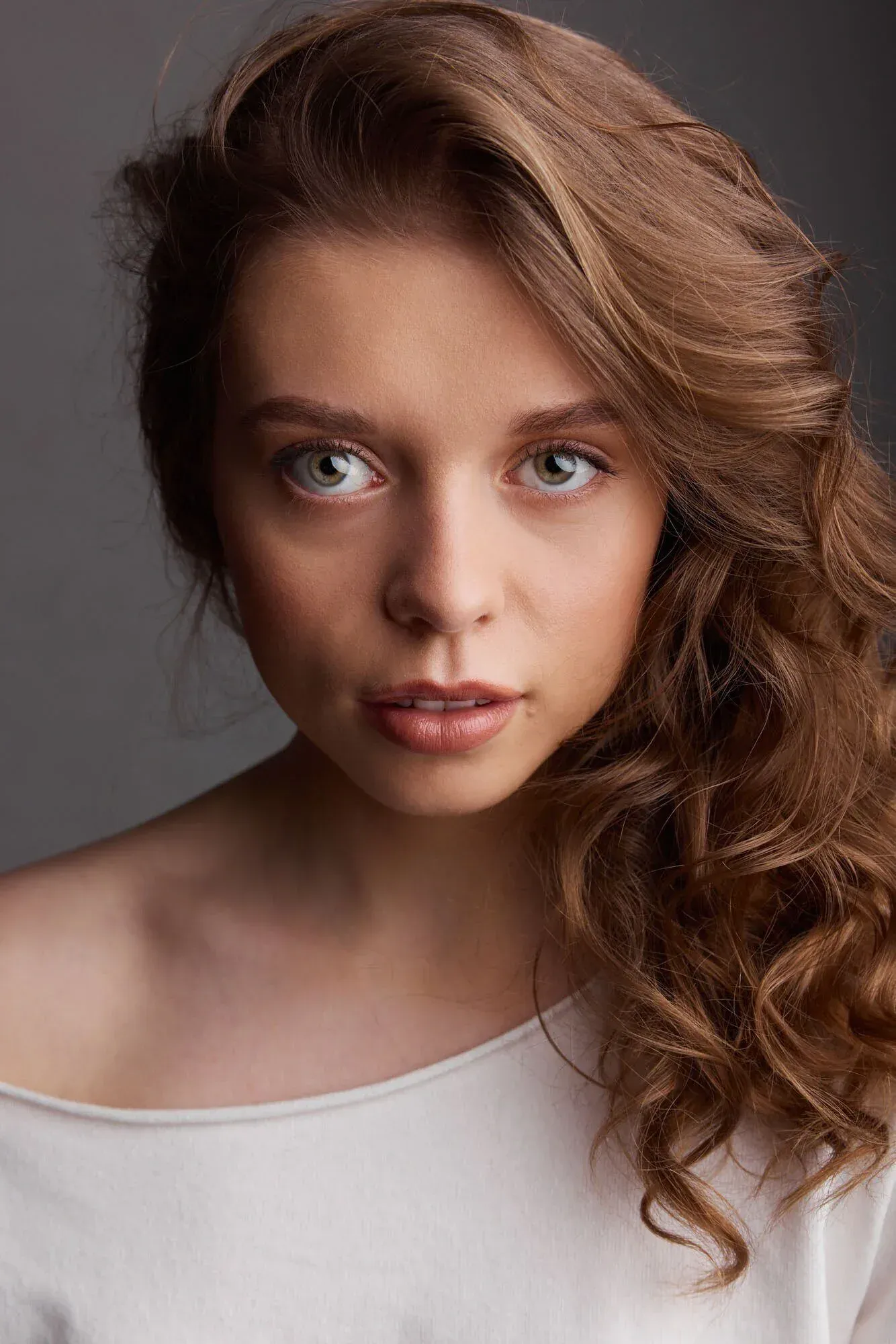 Portrait en gros plan d'une jeune femme aux cheveux auburn bouclés et aux yeux bleus, vêtue d'un haut blanc à épaules dénudées, sur fond gris.
