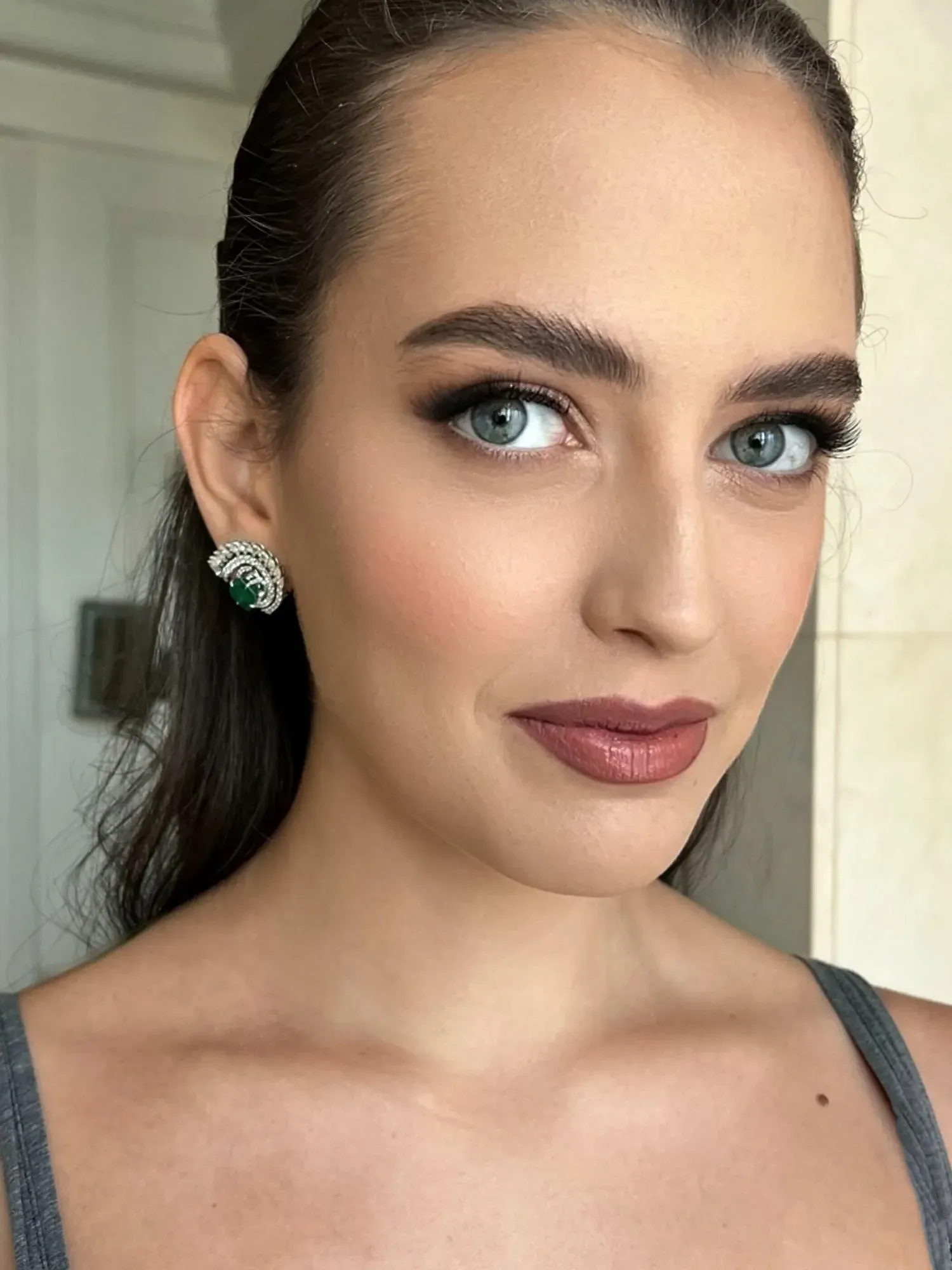Gros plan sur le visage d'une femme, mettant l'accent sur ses yeux bleus, ses sourcils, son maquillage et sa boucle d'oreille ornée d'une pierre précieuse verte.