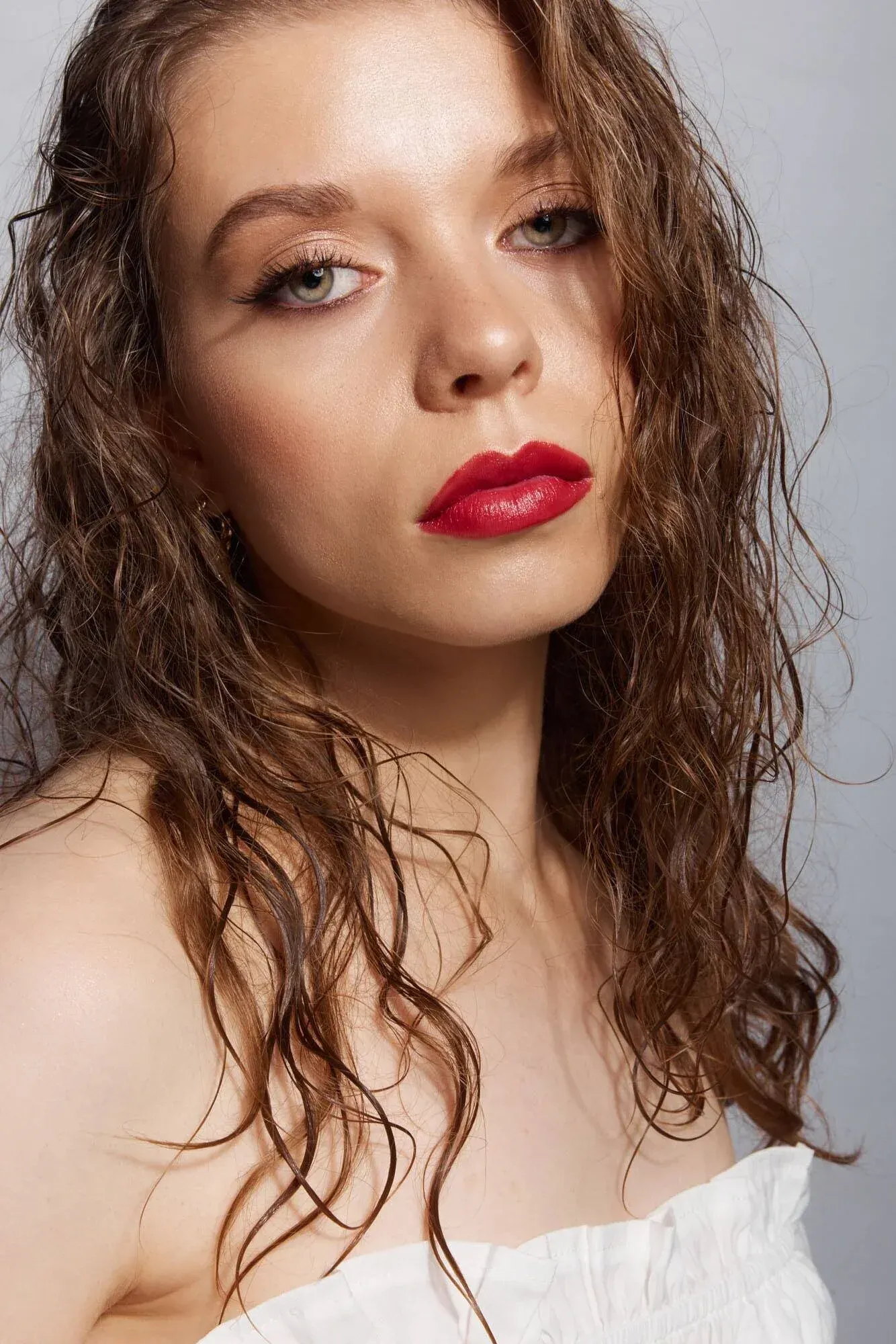 Une jeune femme aux cheveux bouclés auburn et aux yeux bleus, maquillée et portant du rouge à lèvres, qui regarde l'appareil photo sur un fond neutre.