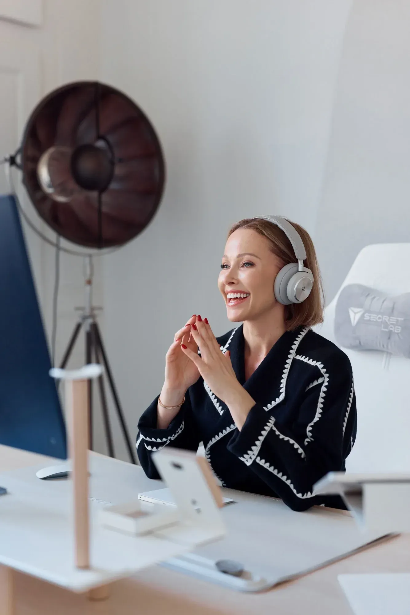 Une femme aux cheveux châtain clair et au rouge à lèvres rouge, portant un pull noir avec des bords brodés blancs, souriant et assise à un bureau avec un ordinateur et un smartphone, portant des écouteurs gris, dans une pièce bien éclairée avec une grande lampe de studio sur une table.