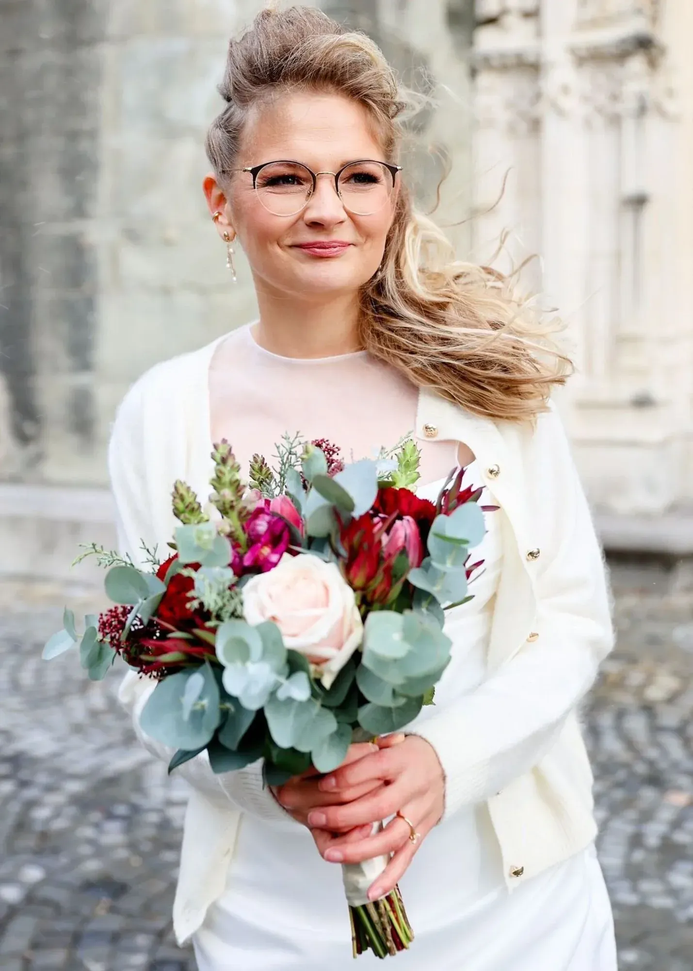 Une femme à lunettes tenant un bouquet de fleurs roses, rouges et blanches, agrémenté de feuillage vert, dans une rue pavée.