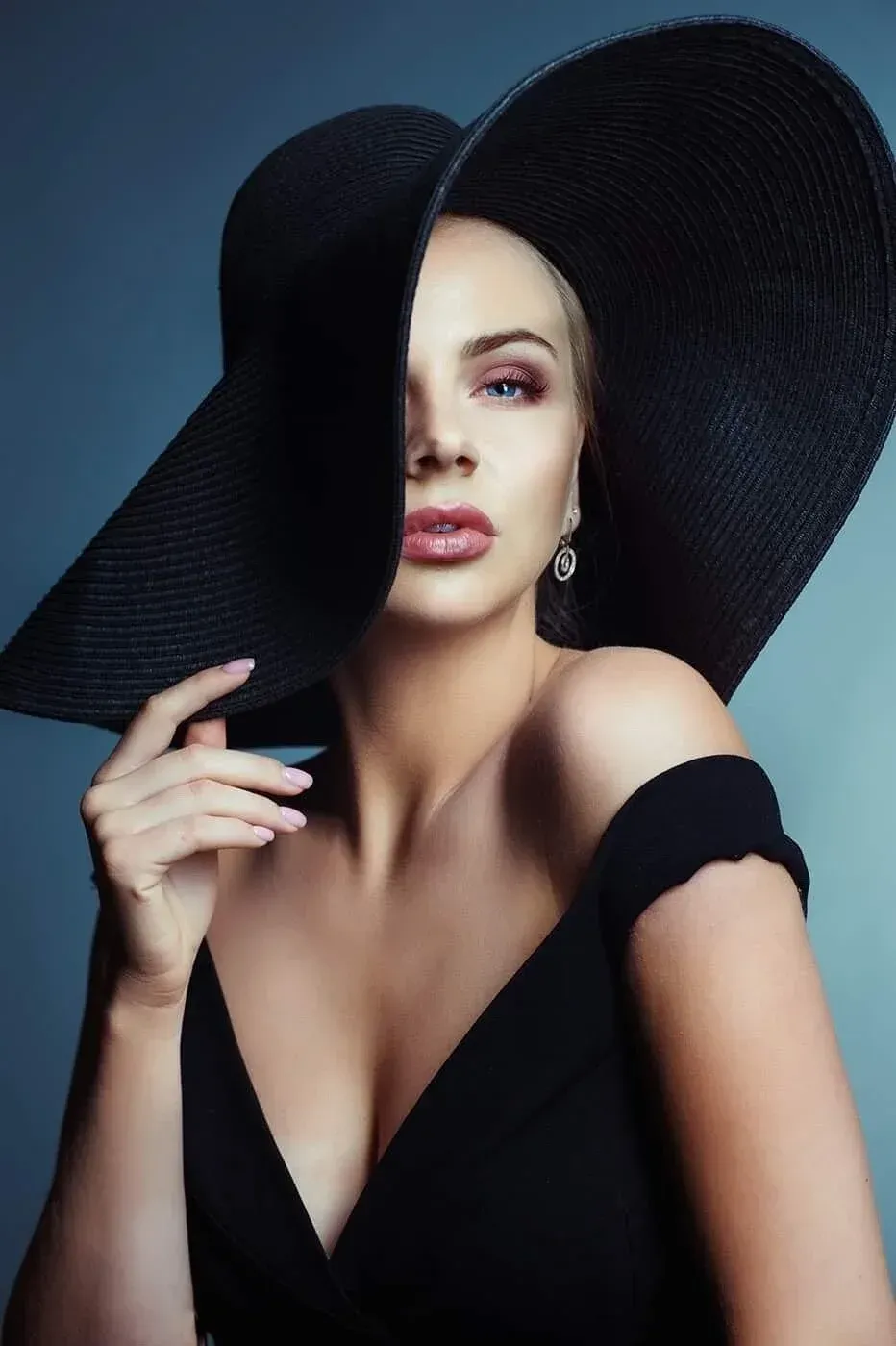 Une femme portant un grand chapeau de soleil noir et un haut noir décolleté, avec un œil légèrement fermé et des lèvres légèrement entrouvertes.