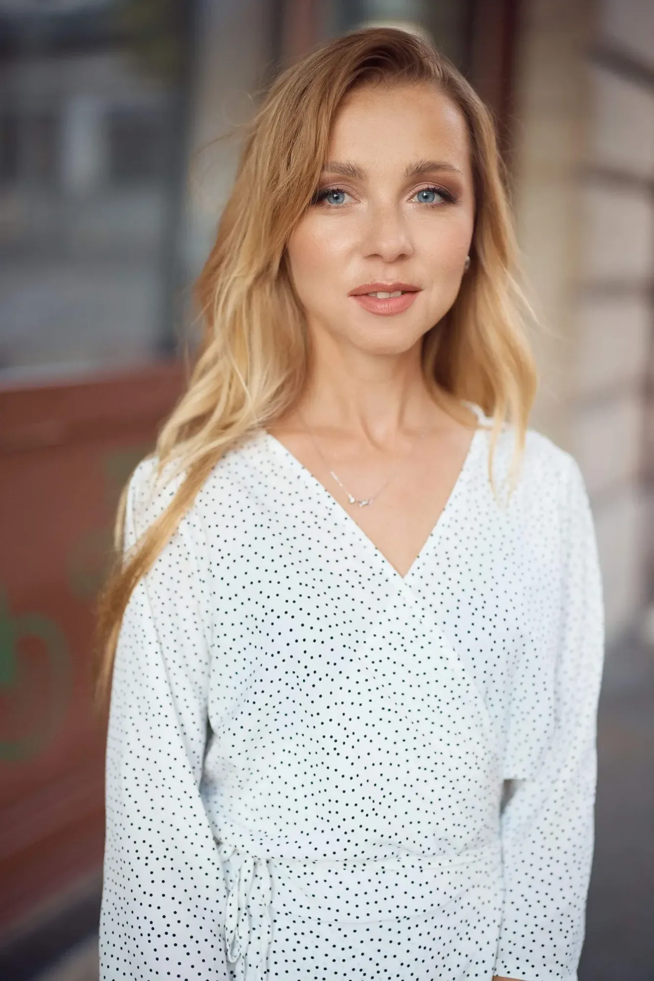 Une jeune femme aux cheveux blonds ondulés et aux yeux bleus, portant une robe blanche à pois et un collier délicat, se tient à l'extérieur avec un léger sourire.