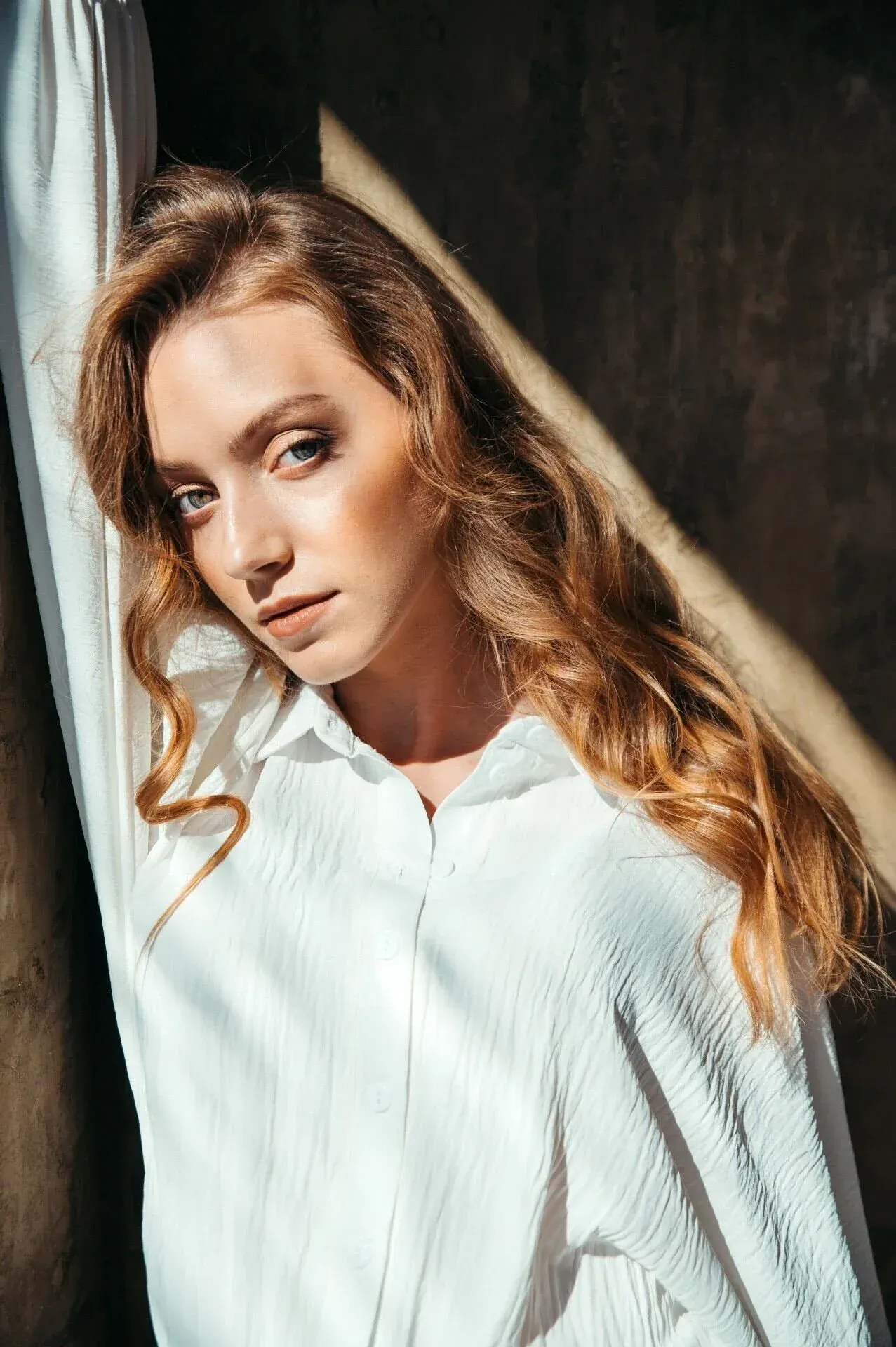 Une jeune femme aux cheveux ondulés et auburn, aux yeux bleus, vêtue d'un chemisier blanc, adossée à un mur traversé par la lumière du soleil.